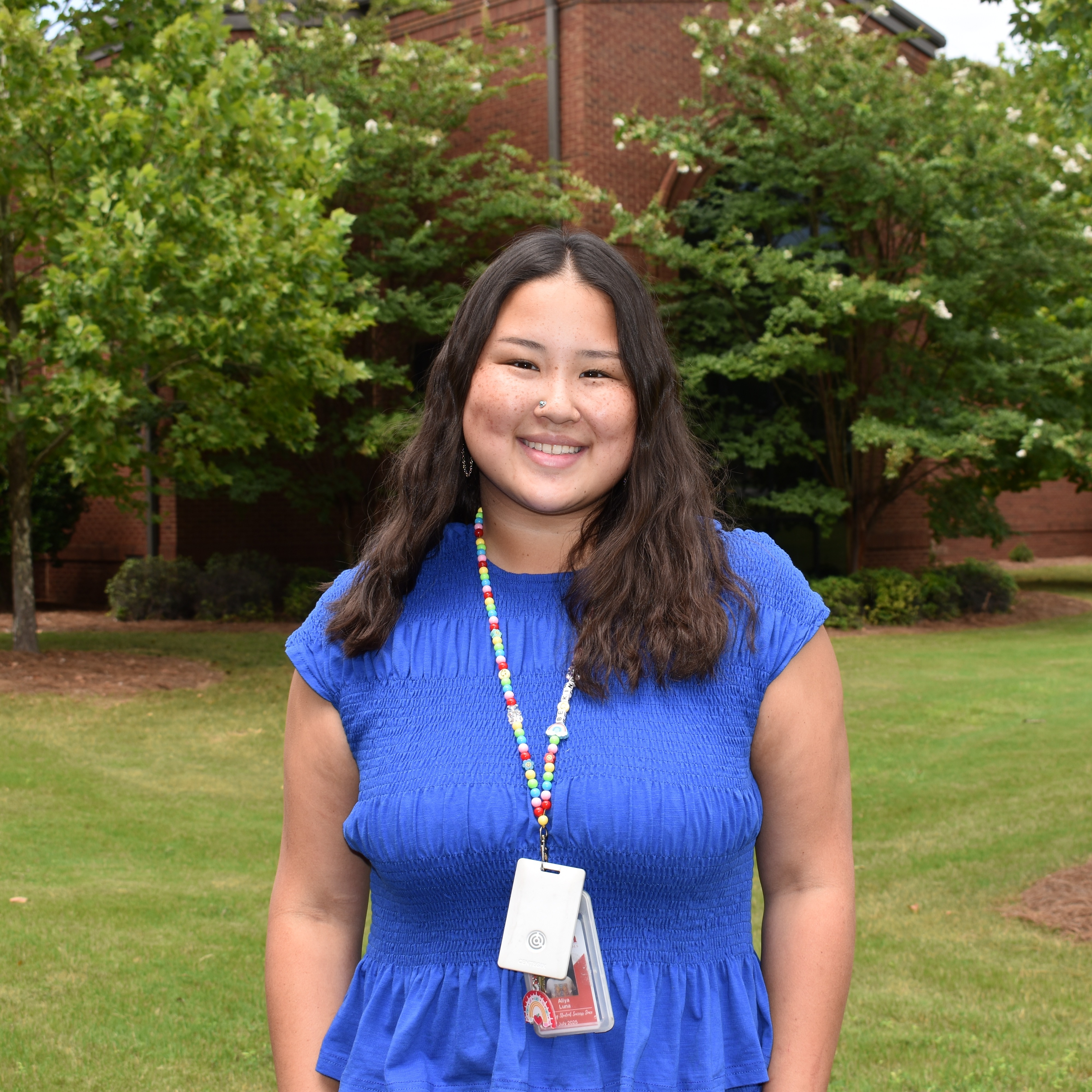 Female with dark hair and blue shirt standing outside in front of trees and a brick building smiling
