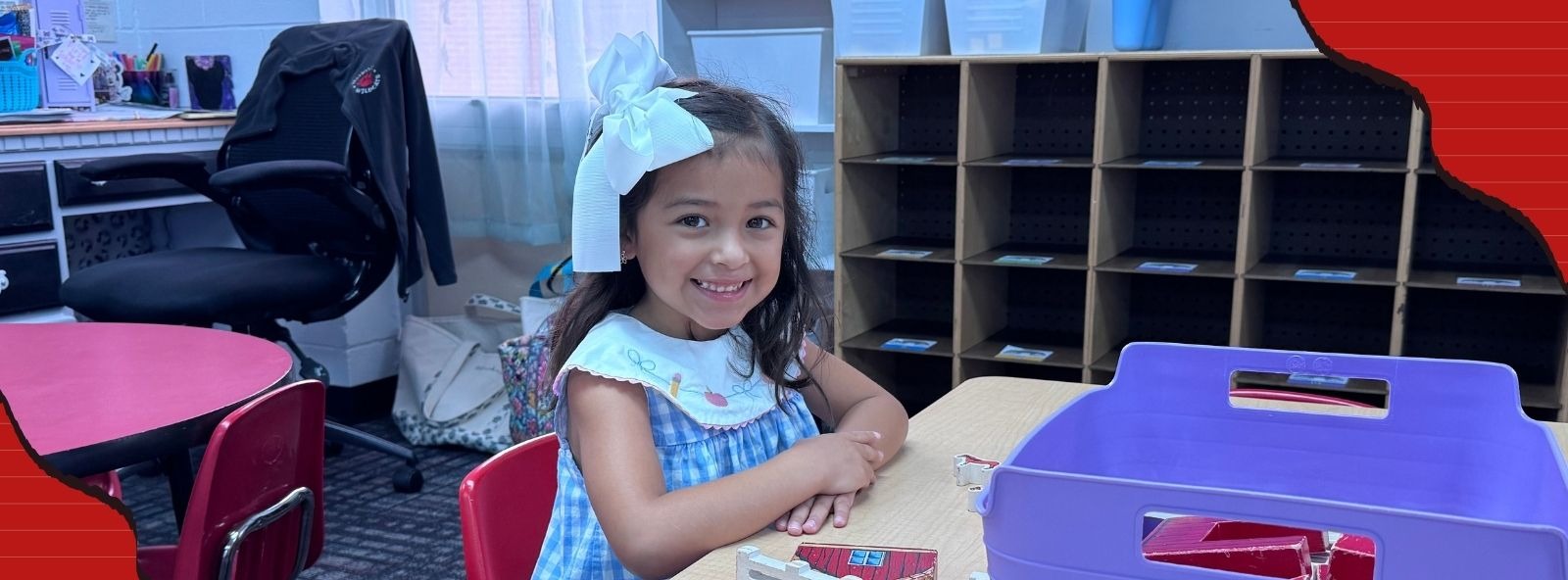 A young girl wearing a blue gingham dress and a large white hair bow sitting at a wooden classroom table, smiling at the camera next to wooden farm blocks and a purple storage bin.