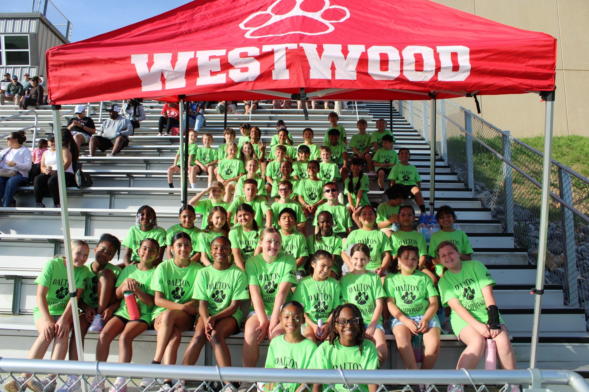 A large group of elementary student athletes wearing matching bright green t-shirts gathered together on stadium bleachers. They are posing for a team photo under a large red tent canopy bearing the name "WESTWOOD" and a paw print logo.