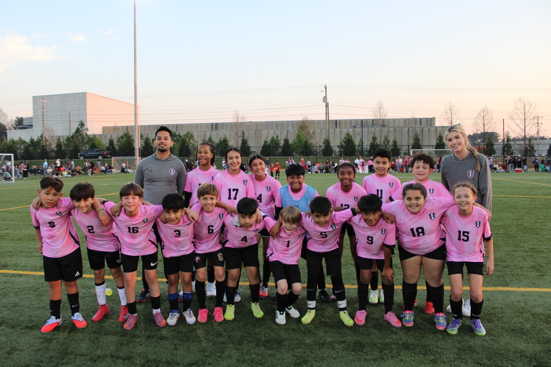 A youth soccer team and their coaches posing together on an outdoor turf field. The players are lined up side-by-side wearing matching pink jerseys with black shorts, with two adult coaches standing behind the team at each end of the row.