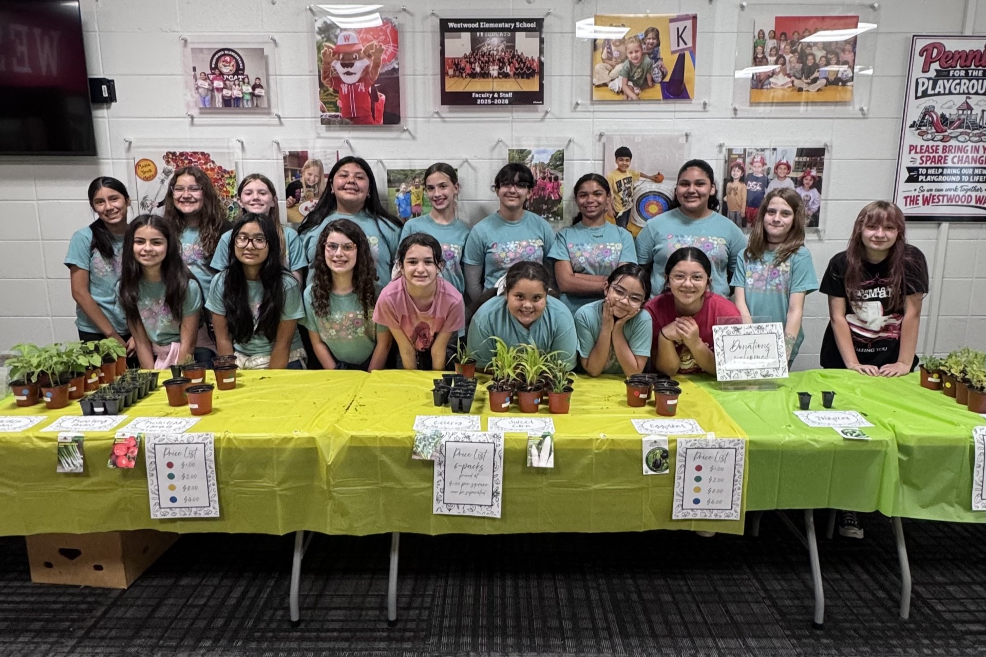 A group of students standing behind tables covered in green tablecloths for a plant sale inside a school hallway. Small potted plants, seedlings, and price lists are arranged along the tables, with school photos and posters displayed on the wall behind them.