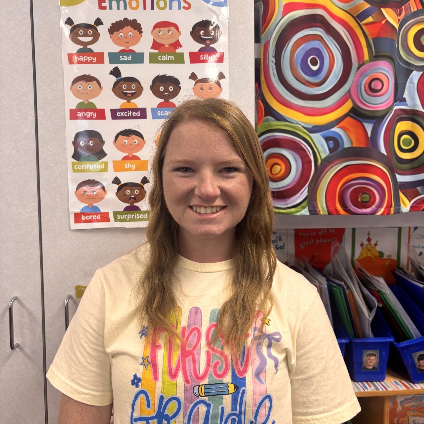 A portrait of a smiling woman with wavy light brown hair wearing a light yellow t-shirt that says "First Grade" in colorful lettering. She is standing inside a classroom in front of an educational poster illustrating various emotions and a colorful, concentric-circle patterned curtain.