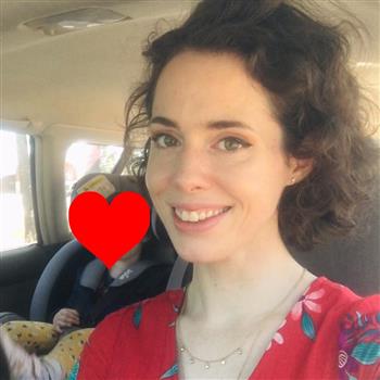 A woman with curly hair sitting in the back seat of a car, with a red heart-shaped object behind her.