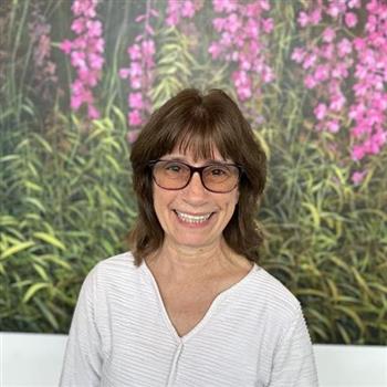 A woman with glasses stands in front of a painting of flowers, wearing a white shirt.