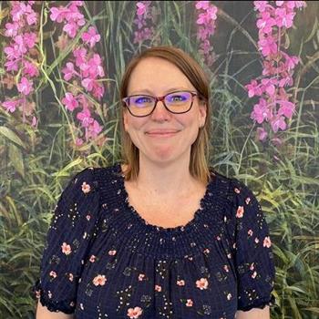 A woman with glasses stands before a vibrant wall of flowers, creating a beautiful and colorful backdrop.