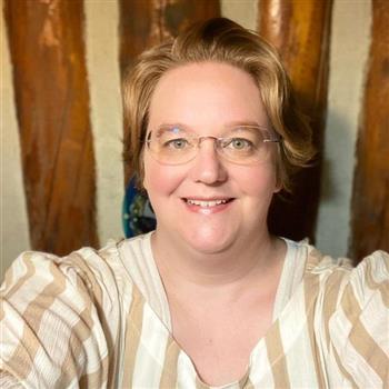 A woman with glasses and a striped shirt standing in front of a bookshelf.