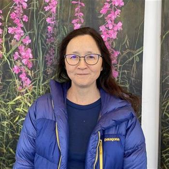 Woman in glasses and blue jacket standing in front of painting.