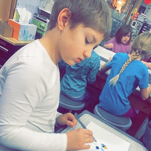 A focused boy sitting at a desk, working on a project in a classroom filled with books and school supplies.