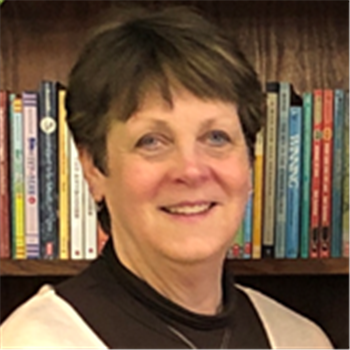 A woman in a black shirt and brown jacket standing in front of a bookcase.