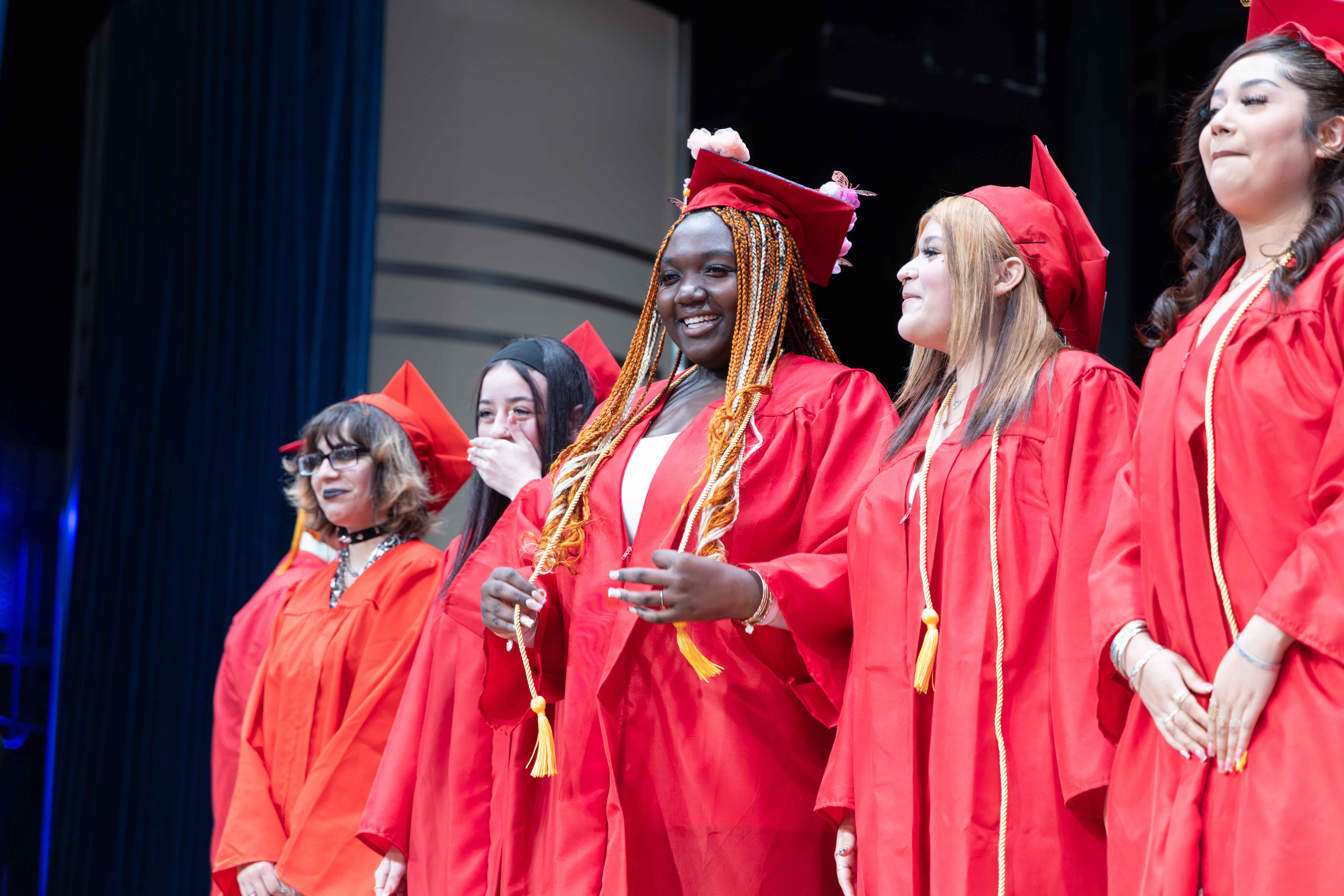 Five women in red graduation gowns and hats, standing in a row on a stage.