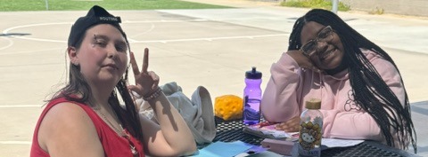 Two women in casual clothing sit at a table outdoors. One woman makes a peace sign. A basketball court is in the background.
