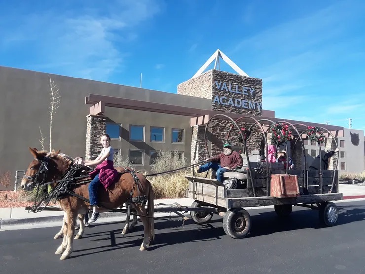 A woman riding a horse-drawn wagon through a parking lot in front of the Vale Academy building.
