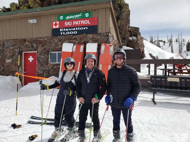 Three people posing for a picture with ski equipment, standing in front of a mountain resort ski patrol station.