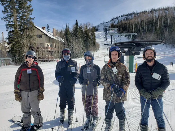 A group of five people, likely skiers or snowboarders, dressed in winter gear, posing for a photo on a ski slope with the sun shining brightly.