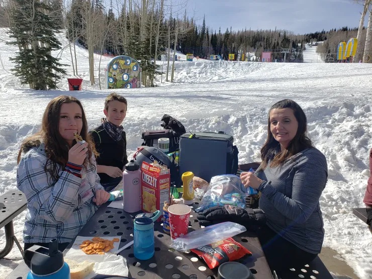 Two adults and a child seated at an outdoor picnic table, enjoying refreshments in a snowy park setting.