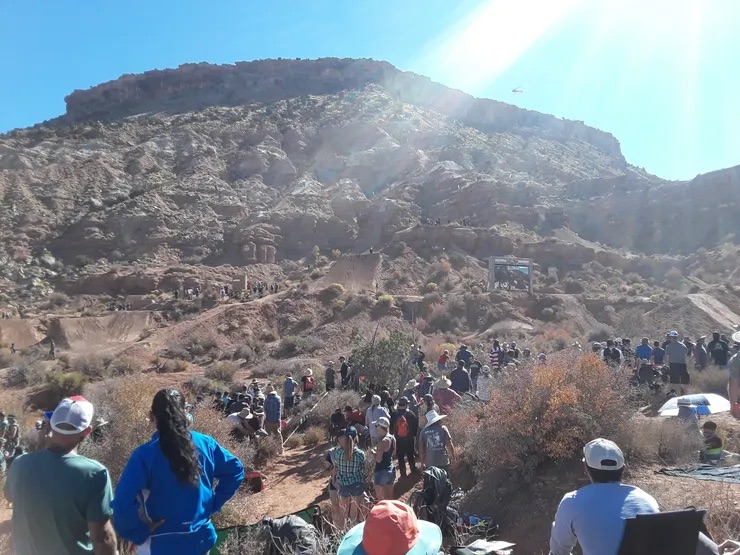 This is a photo of people gathering for an outdoor event, possibly at a sports game or concert in the daytime. They are seated on various terrains with a mountain backdrop and clear skies overhead.