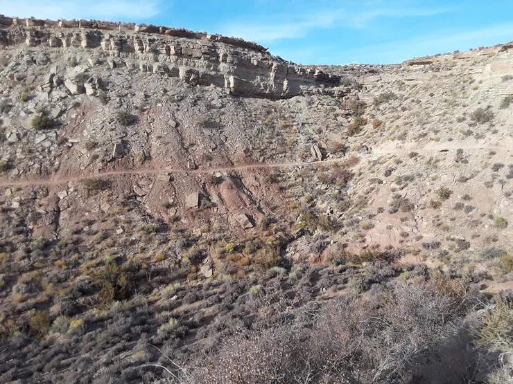 A rugged landscape featuring a dirt road cutting through a barren, rocky hillside with sparse vegetation and a clear blue sky.