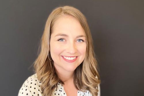 A woman with blonde hair wearing a white polka dot blouse, smiling at the camera against a gray wall.