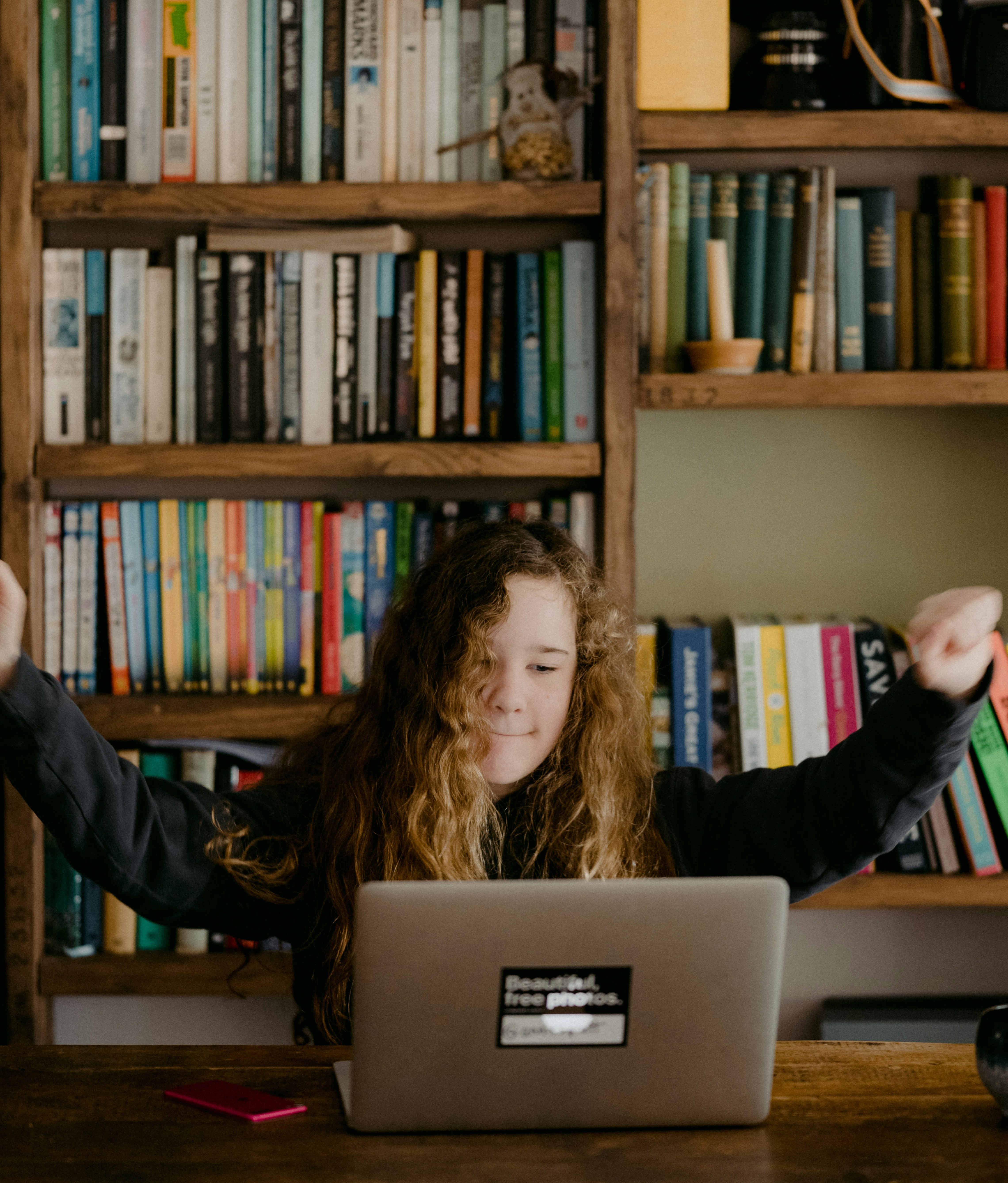 Girl sitting in front a computer pumping her arms in the air with a bookcase of books behind her