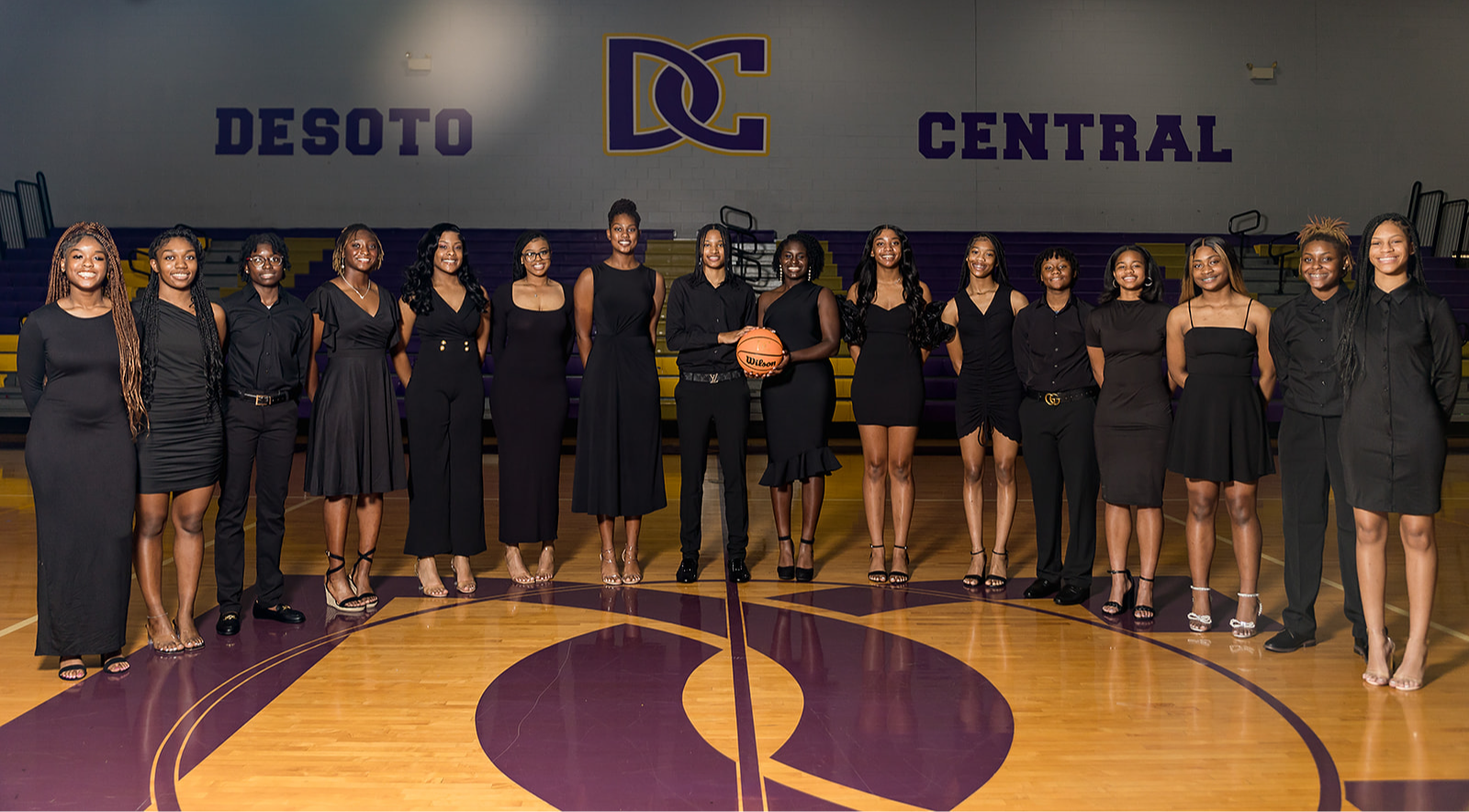 DCHS Lady Jags basketball team wearing black business semi-formal clothes, standing in a semicircle on the basketball court