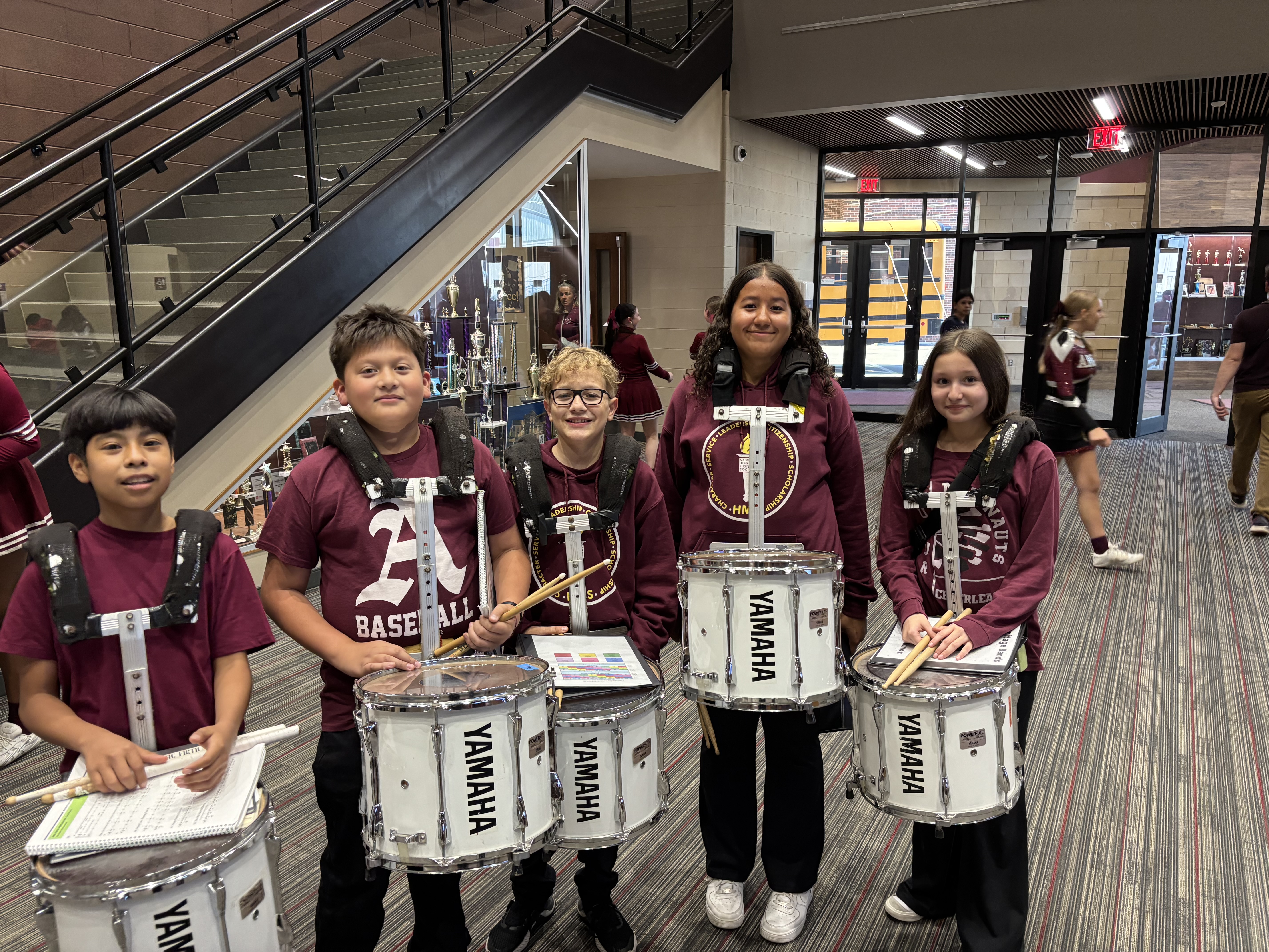 Five young students in maroon outfits hold snare drums and drumsticks in a school hallway, smiling near stairs, with cheerleaders in the background.