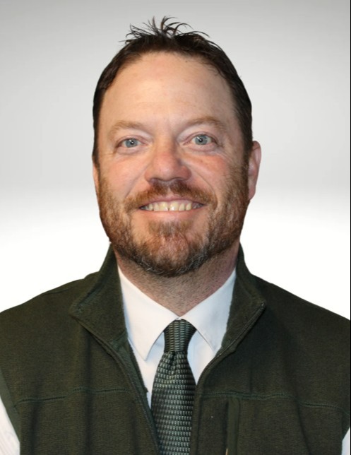 Smiling man with a brown beard and short hair, wearing a green vest over a white shirt and dark tie, against a light gray background.