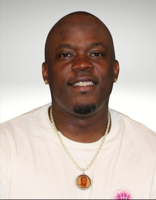 Smiling man in a white shirt with a floral design and a gold chain necklace. The background is plain, conveying a friendly and relaxed tone.
