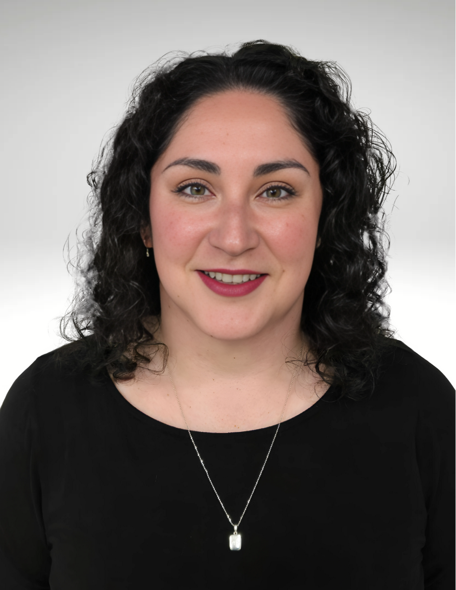 Smiling woman with curly hair wears a black top and a silver necklace. She stands against a neutral background, conveying a friendly and approachable demeanor.