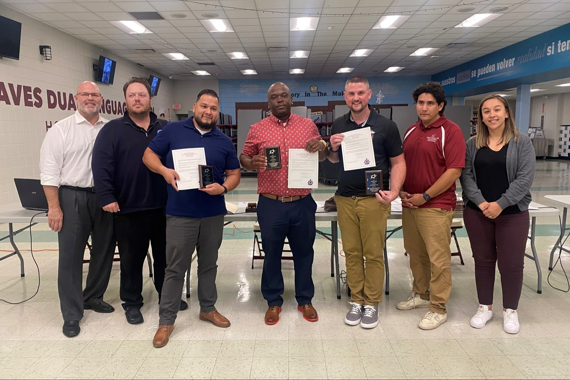 A group of seven people stand indoors, three holding certificates and awards. The setting is a classroom or cafeteria with a positive, celebratory atmosphere.