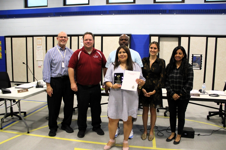 A group of six people standing in a room, with a central woman holding a plaque and certificate, all smiling. The setting appears to be professional.