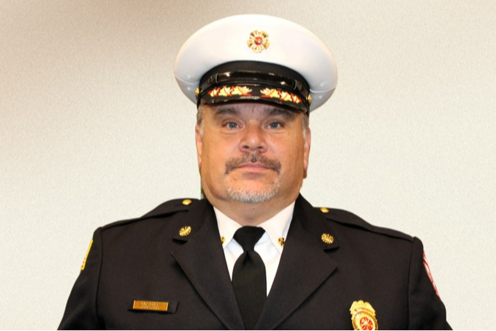 A fire chief in a formal uniform with badges stands against a neutral background, conveying a sense of authority and pride. He holds a wrapped gift.