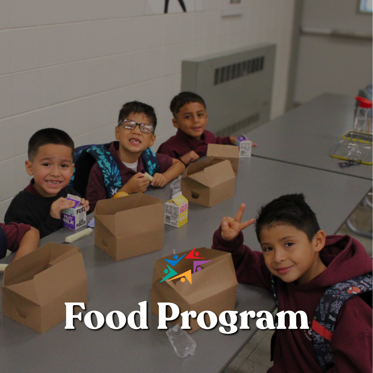 Four smiling schoolchildren sit at a classroom table with food boxes and milk cartons; one flashes a peace sign beneath the words “Food Program.”