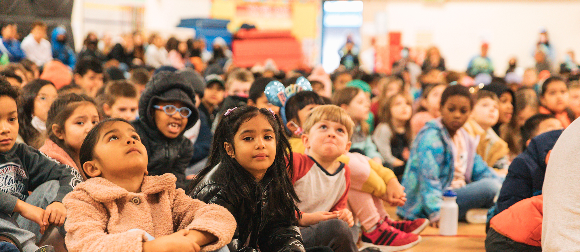 A group of children sit on a gym floor attentively watching something in front of them