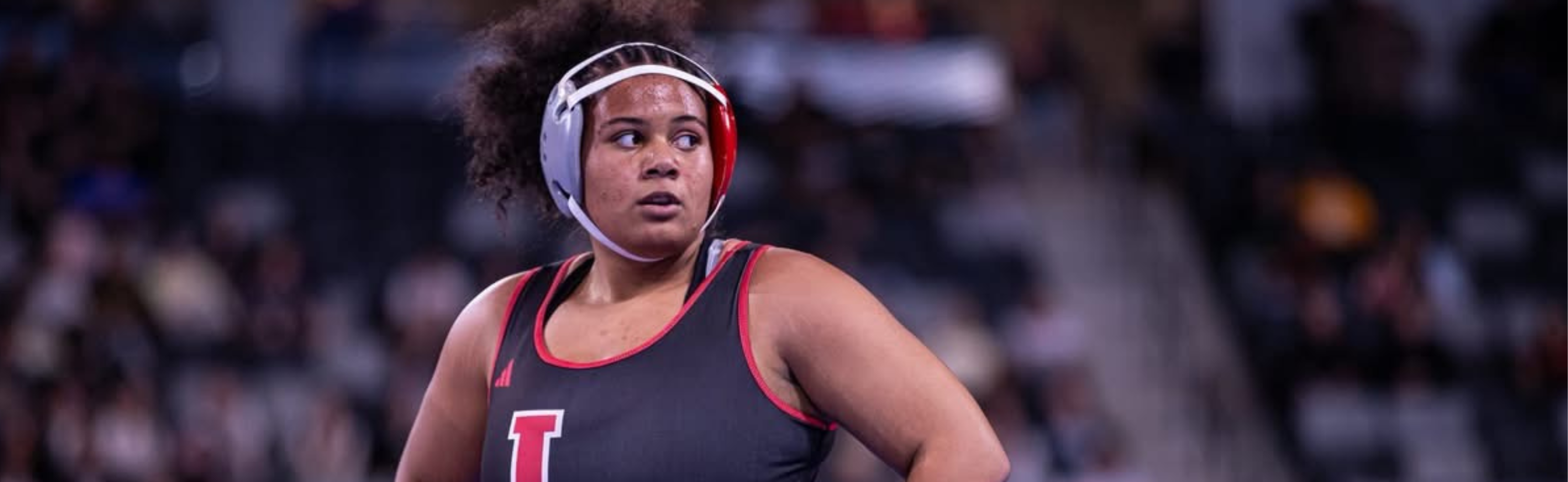 A female athlete wearing a wrestling uniform stands on a court, looking to the side.