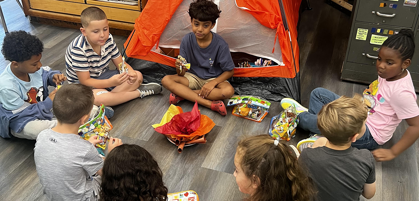 students sitting outside a tent around a make believe campfire