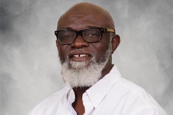 A man with a beard, glasses, and a white shirt stands smiling, with a flag in the background.