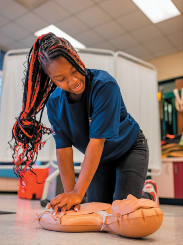 A person practices CPR chest compressions on a training mannequin in an indoor classroom setting.