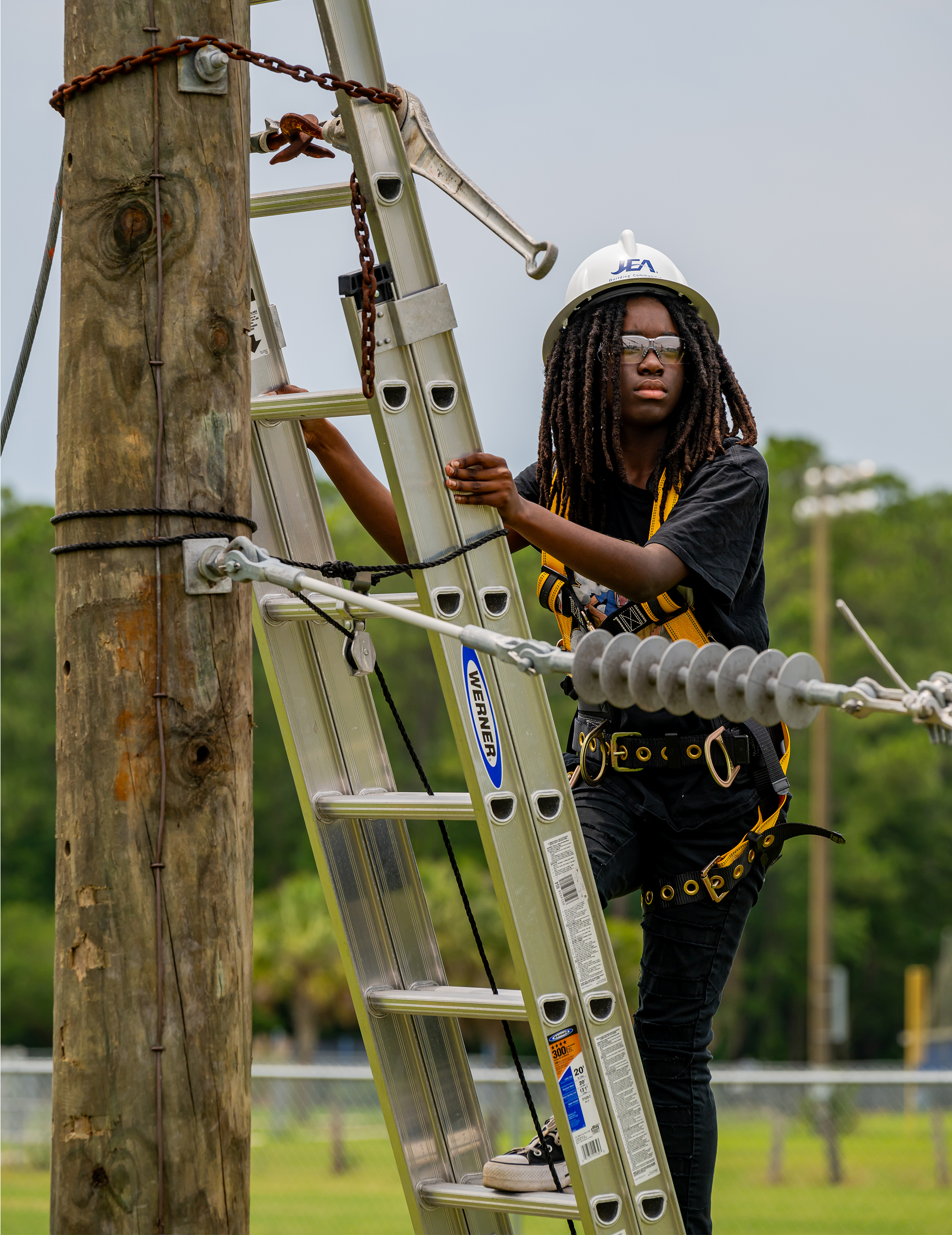 A person in protective gear ascends a ladder on a utility pole outdoors, focused and determined. They wear a safety harness and hard hat.