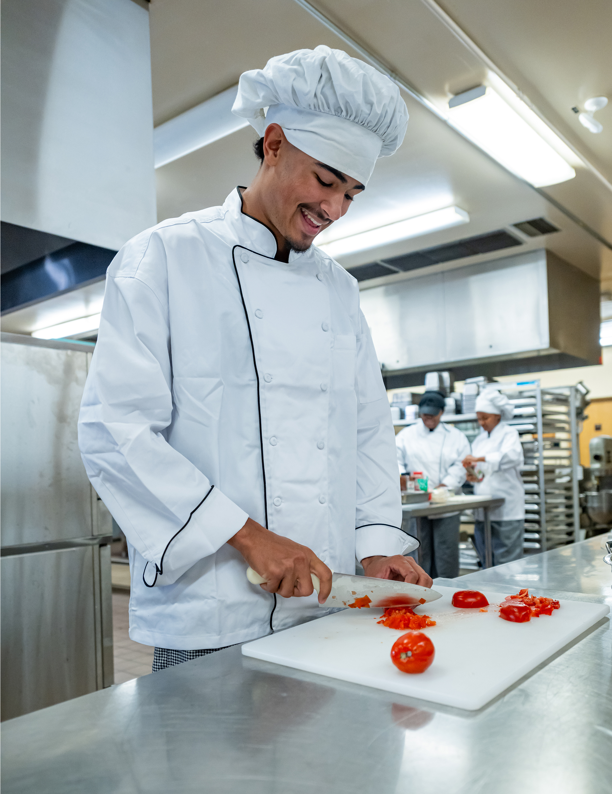 A chef in a white uniform and hat smiles while chopping tomatoes on a cutting board in a professional kitchen. Two other chefs work in the background.