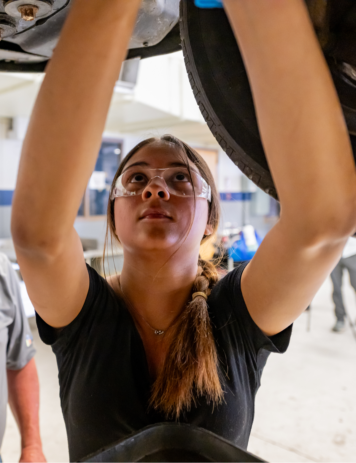 A young woman with safety goggles works intently beneath a vehicle in a bright workshop, conveying focus and dedication in a hands-on setting.