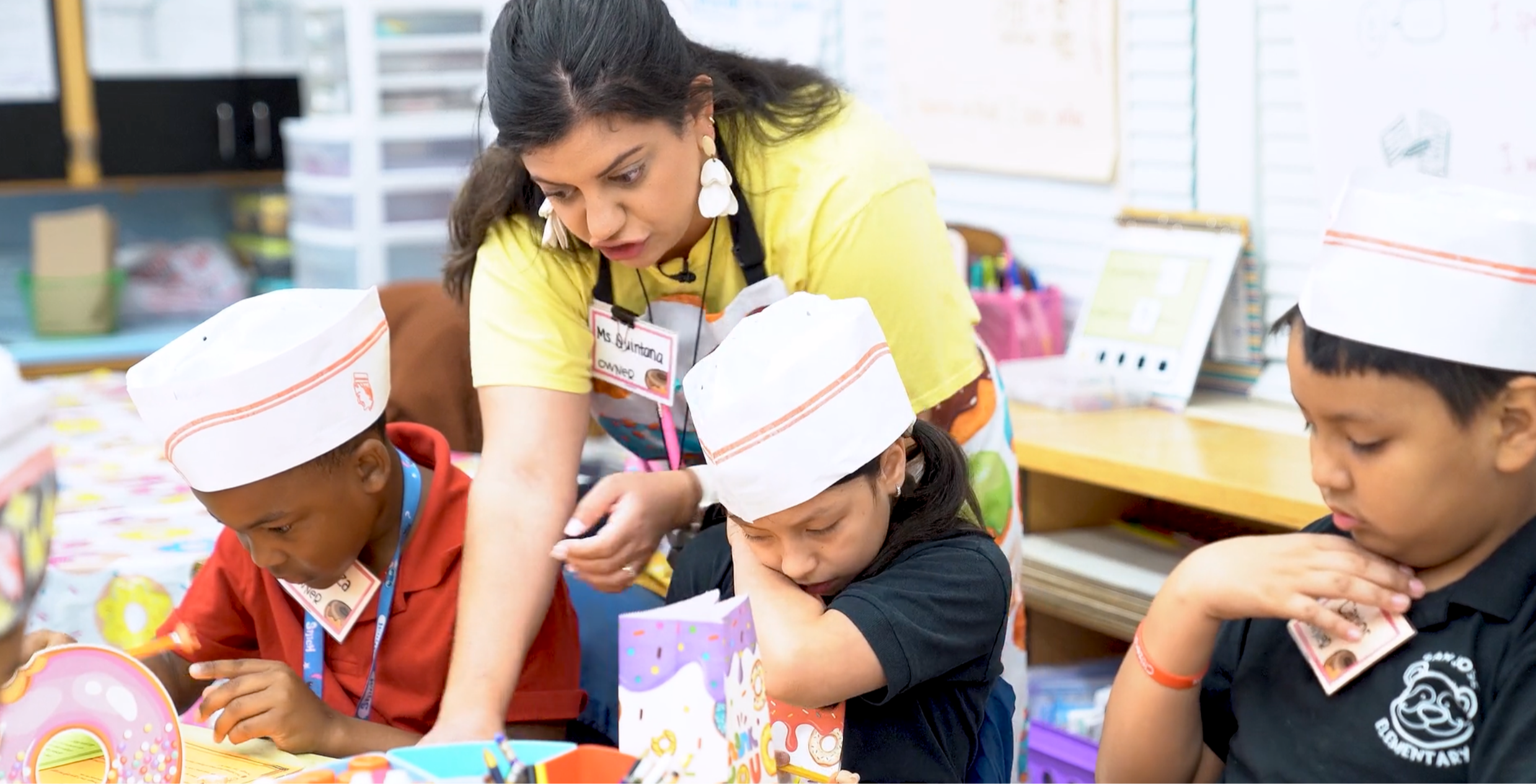 Teacher Alex Quintana helps second-grade students wearing paper donut shop hats during a hands-on reading activity at San Jose Elementary.