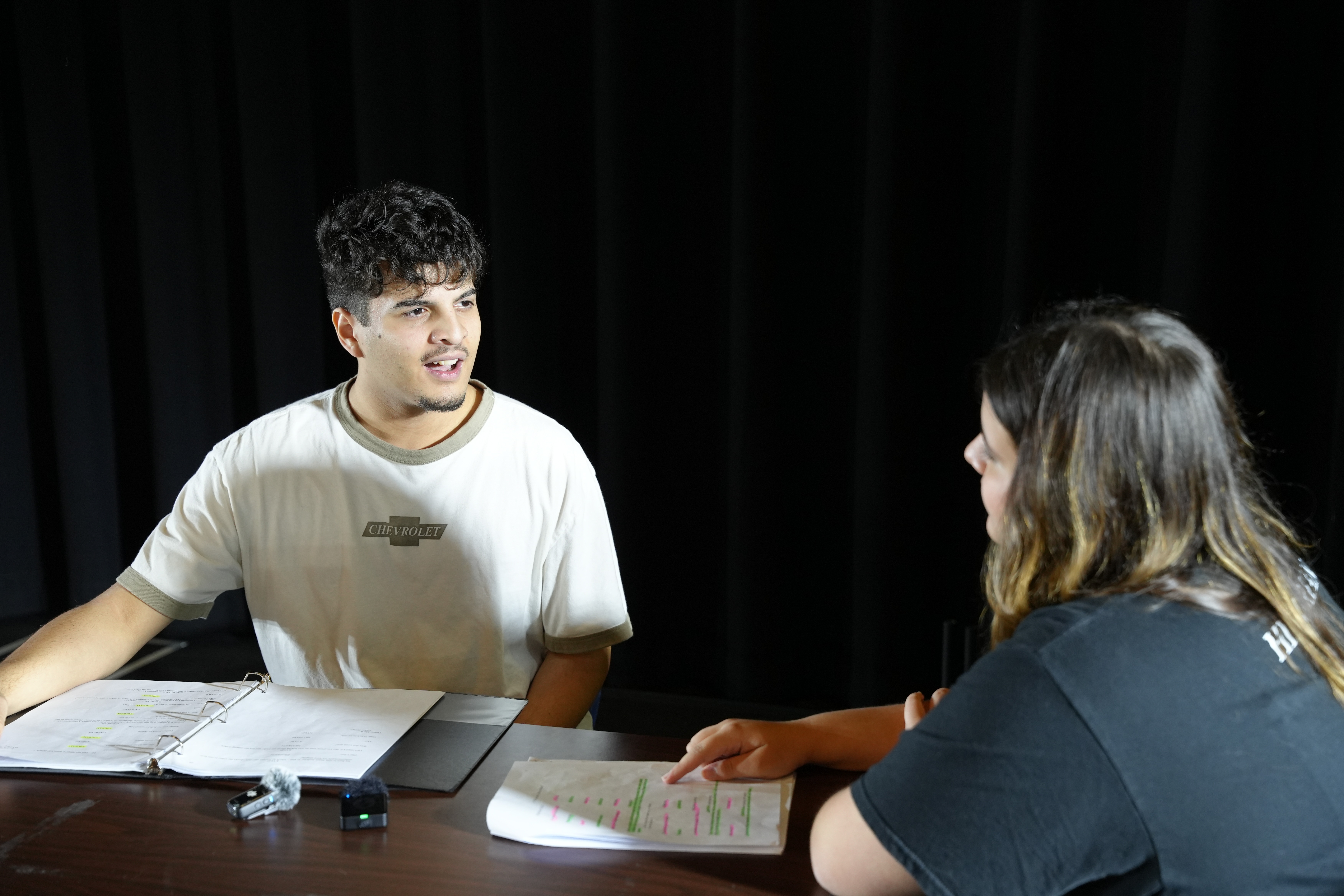Two Wolfson High School theatre students rehearse a scene together under stage lighting, reviewing scripts and practicing lines for an upcoming production.