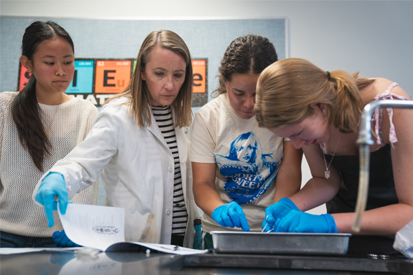 A science teacher in a white lab coat guides three students wearing blue gloves as they perform a dissection in a biology lab.