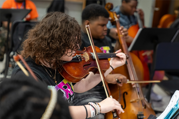 A close-up of a student playing the viola in an orchestra class, with other students playing cellos in the background.