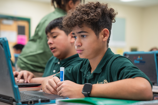 A student wearing a green polo shirt focuses intently on a laptop screen while holding a marker, sitting next to a classmate.