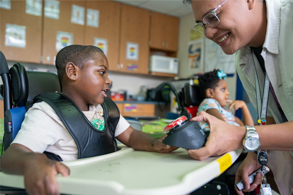 A smiling educator holds a red adaptive switch button for a young student in a wheelchair who is reaching out to press it.