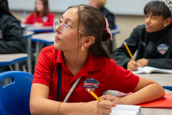 A female student wearing glasses and a red Joseph Stilwell Military Academy polo shirt sits at a desk, looking up attentively with a pencil in hand.