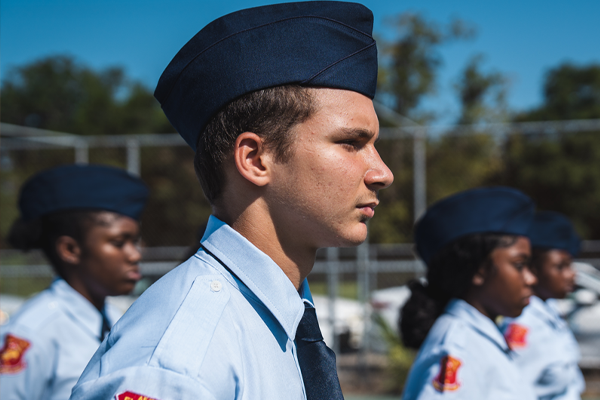 A male student in a light blue military uniform and flight cap stands in profile during an outdoor formation, with other cadets standing in line behind him.