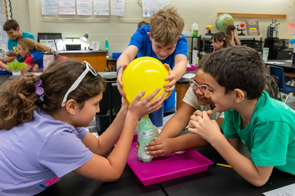 Four young students gathered around a science lab table hold a yellow balloon attached to the top of a plastic bottle containing green liquid.