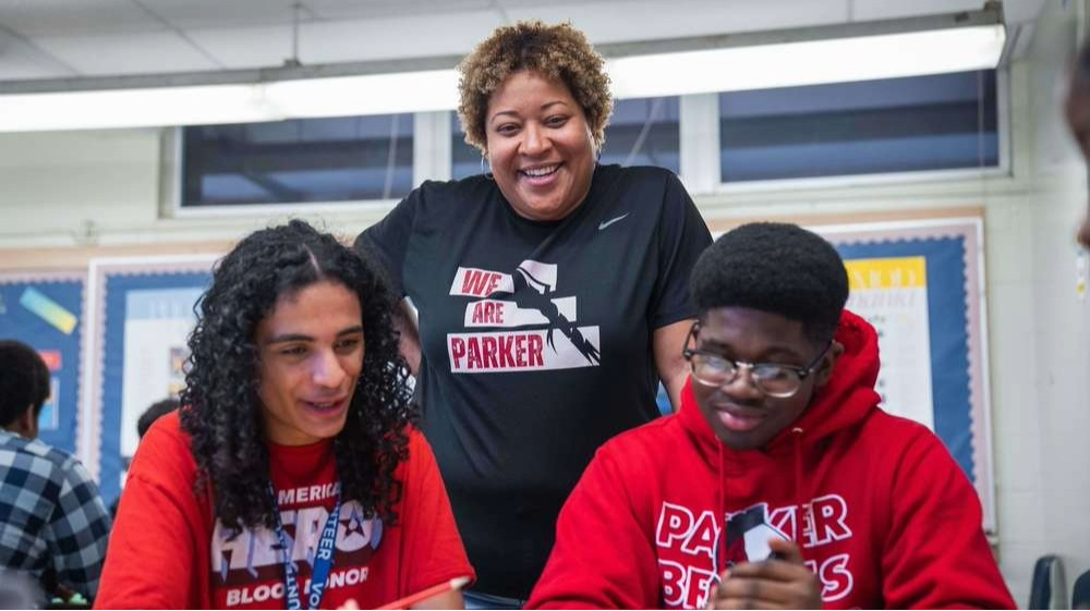 Two students in the classroom smile as they work together on a project. A smiling teacher stands behind them.